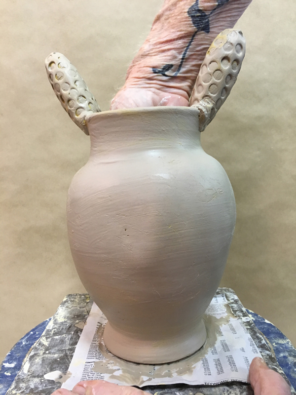 2 Exaggerate the shape by pushing with your fingers. Add decorative handles and brush a white slip over the entire vase.