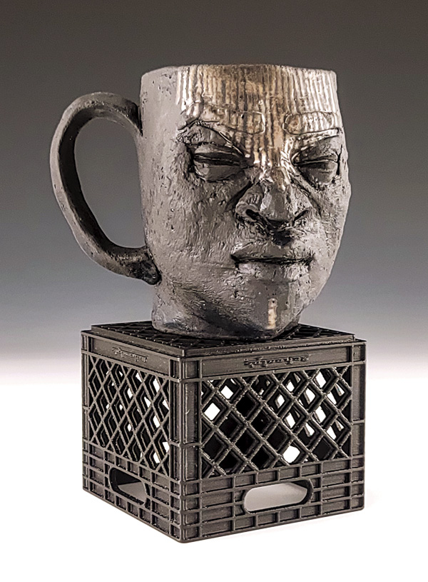 2 Marcè Nixon-Washington’s Ife Steel City Face Mug, to 7¼ in. (18.4 cm) in height, stoneware, gold luster, 3D-printed milk crate, 2025.