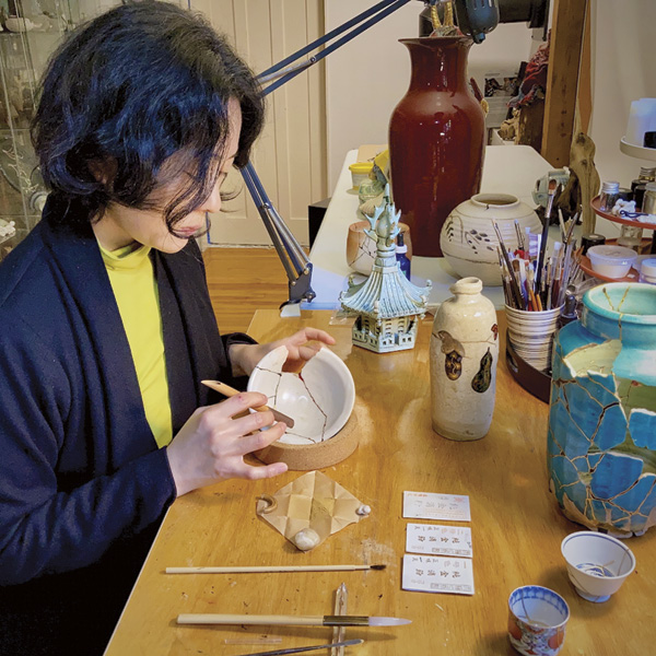 3 Fukumaru in her studio in Vancouver, British Columbia, in 2024 with a funzutsu, a bamboo tube with the end covered in silk, applying gold powder over the top layer of lacquer used in the repair of a bowl.