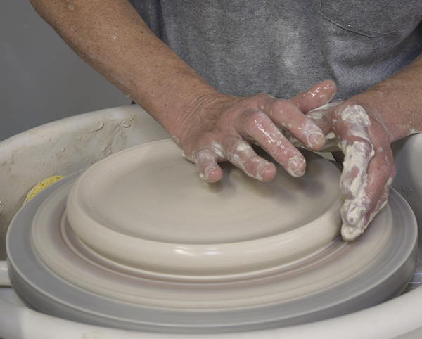 1 To start, center 5 pounds (2.3 kg) of clay and bring it into a pancake about 10–11 inches (25.4–27.9 cm) across. Use the heel of your hand to create a trough around the edge.