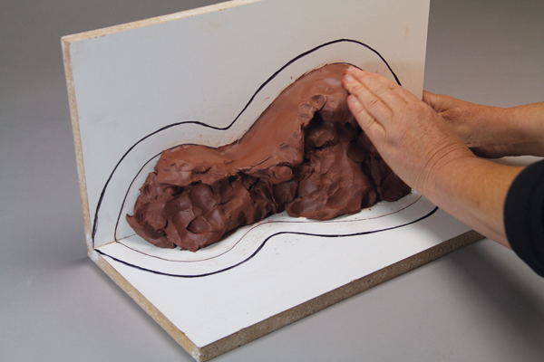 2 Trace the outline of your paper template onto the right-angle support boards. Next draw a line ½ to 1 inch (depending on size of mold) above this outline to mark the thickness of the plaster mold. Begin filling in the space with clay to create the solid clay model.