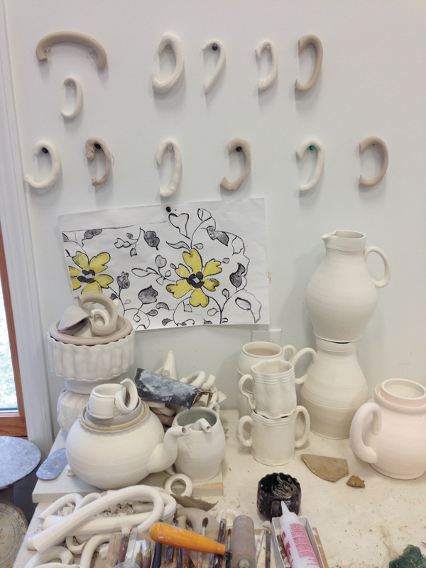 8 Bisque-fired handle shapes and pattern and design ideas hung up on the wall, paper templates, additional handle and spout shapes, and bisque-fired prototypes that Sikora references when designing forms.